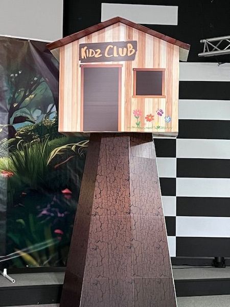 Model of a Kidz Club house sign on a dark brown pedestal, against a backdrop of a jungle scene and black and white squares.