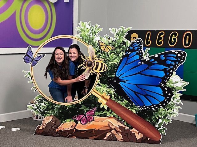 Two women smile in a butterfly-themed cutout, holding hands. The cutout has a magnifying glass and paper butterflies.