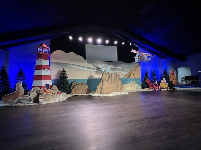 Stage set with a lighthouse, rocks, ocean backdrop, and chairs. Blue and neutral colors dominate.