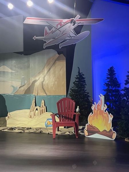 A stage set with a seaplane, chair, campfire, and trees. Painted backdrop depicts a beach, sea, and mountains.