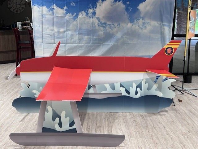 Cardboard cutout of red and white seaplane on blue water, set against a cloudy sky backdrop.