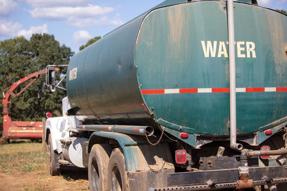Green water tanker truck with a red hose parked outdoors on a dirt field