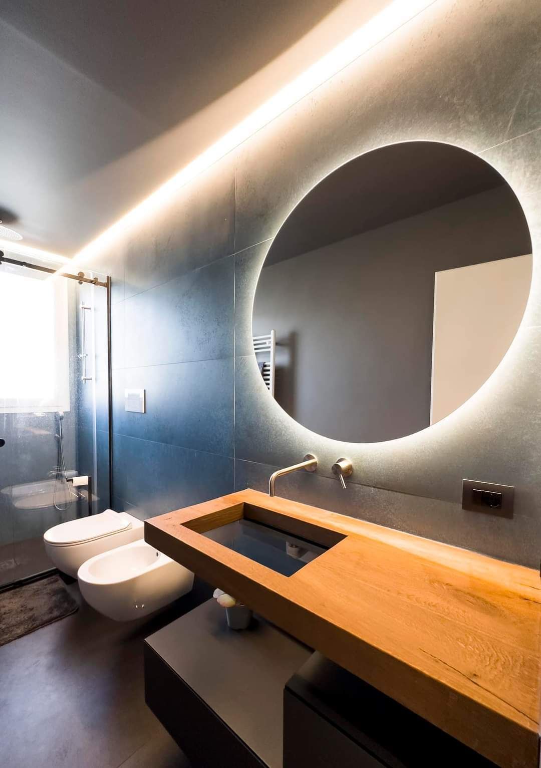 Bagno moderno con pareti grigie, specchio rotondo retroilluminato, mobile lavabo in legno e WC sospeso.