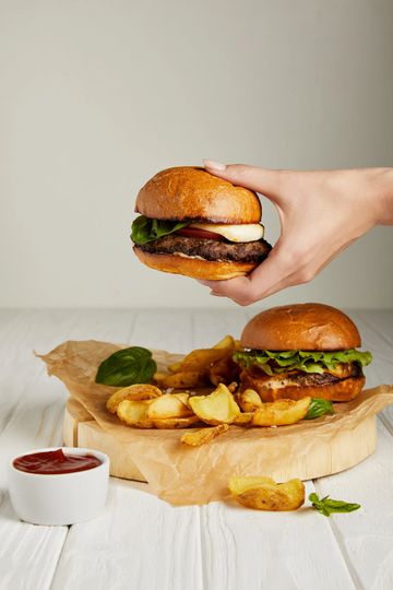 Mano che tiene un hamburger sopra un tagliere di legno con un altro hamburger, patatine fritte e ketchup.