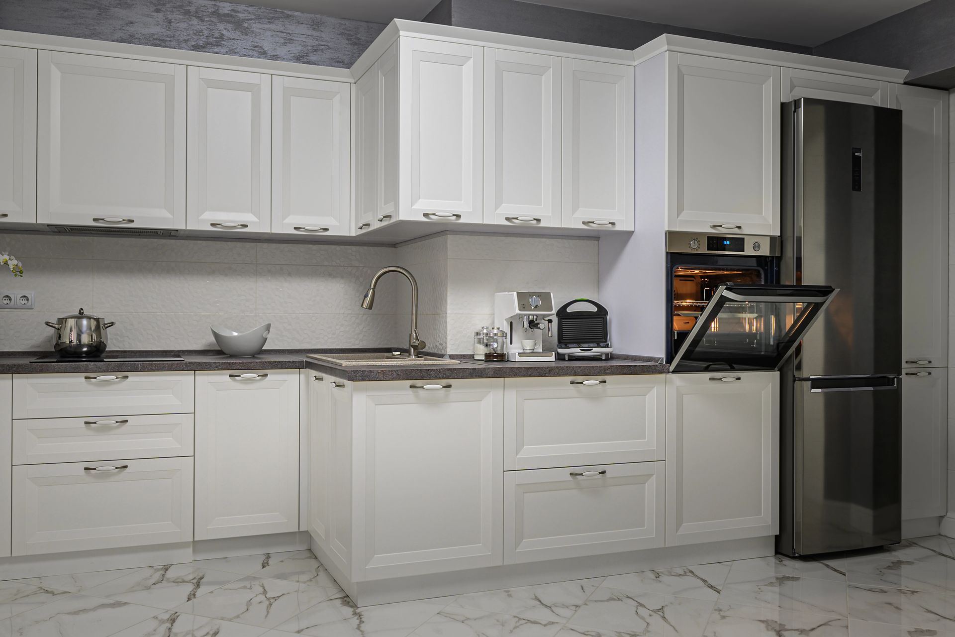 Una cucina moderna con mobili bianchi, ripiani in marmo scuro, frigorifero in acciaio inox e forno a vista.