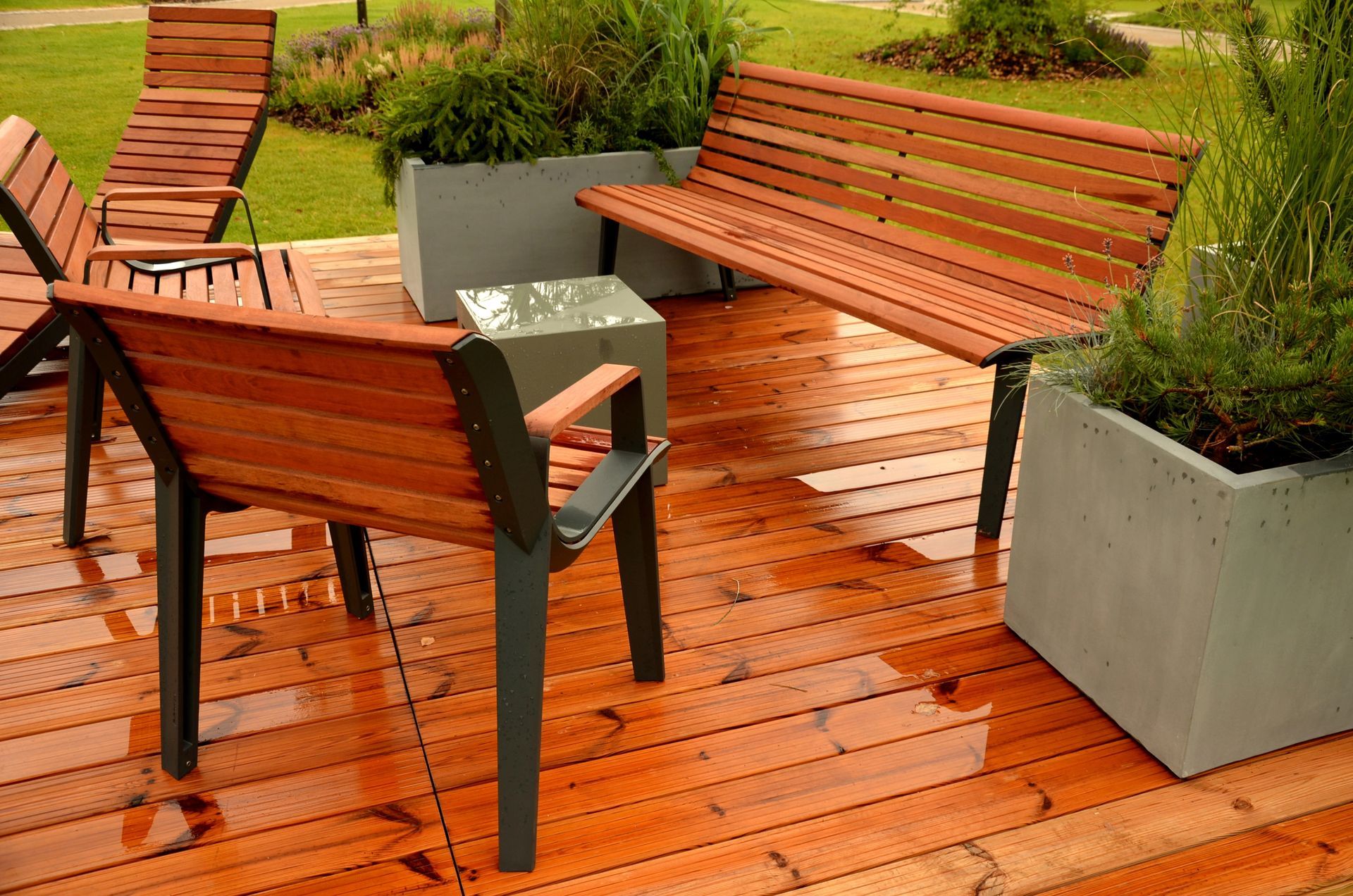Un set da patio con panca, sedia e sdraio in legno su una terrazza in legno bagnata, circondato da fioriere in pietra grigia.