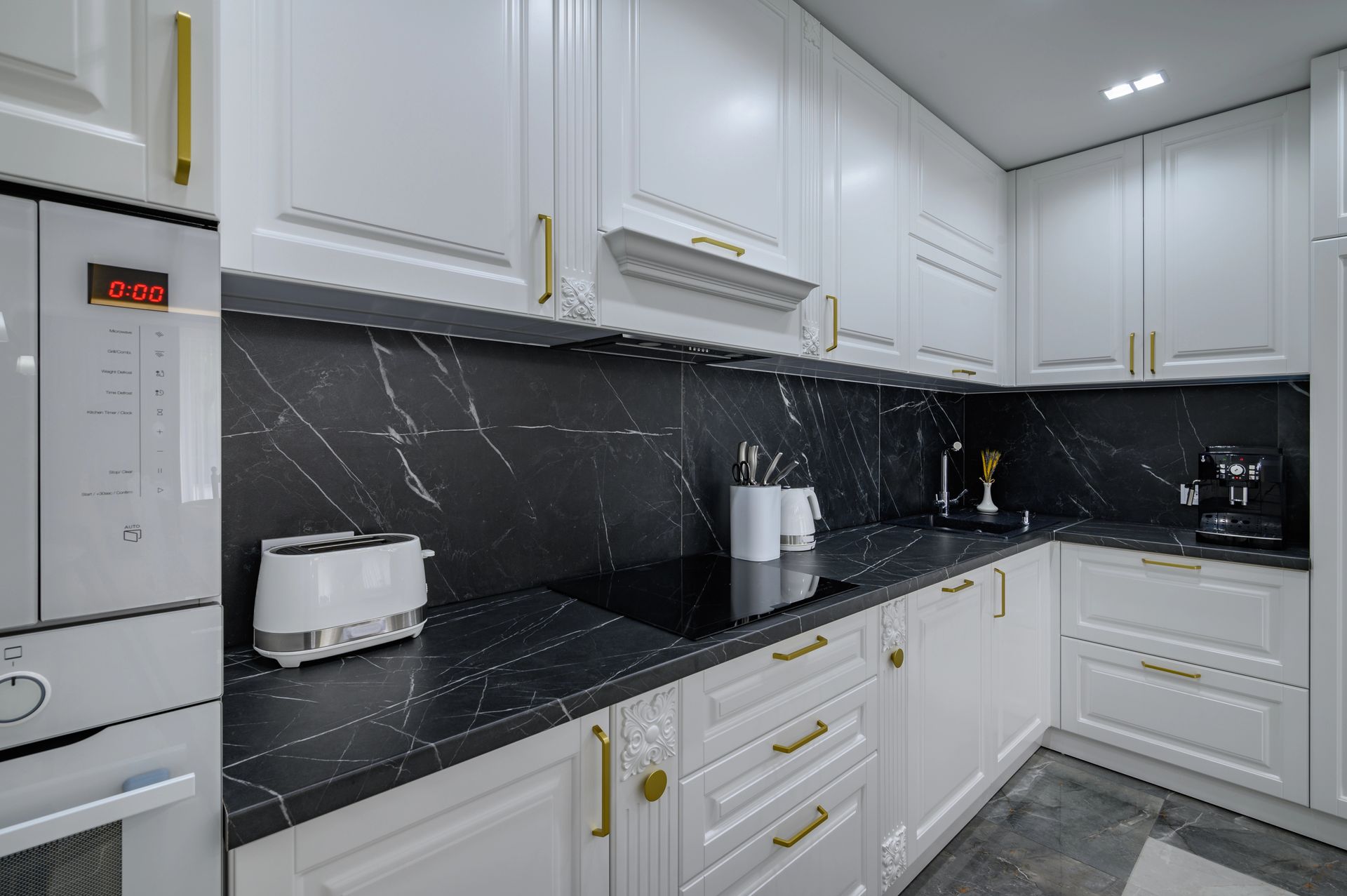 Una cucina moderna con mobili bianchi, maniglie dorate e ripiani e paraschizzi con motivo effetto marmo nero.