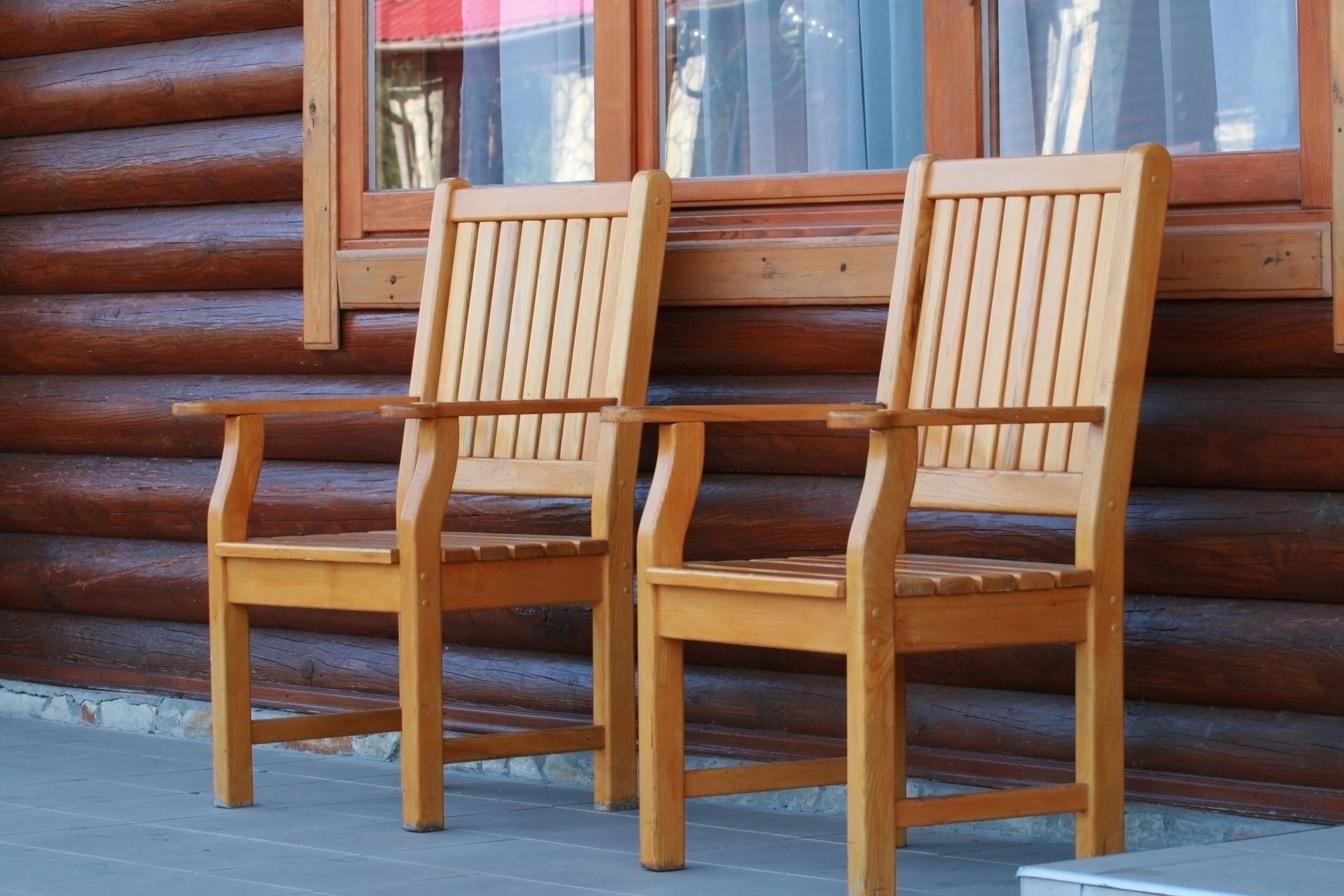 Due poltrone in legno marrone chiaro con schienale a doghe sono posizionate su una terrazza di fronte a una parete e una finestra di una baita di montagna.