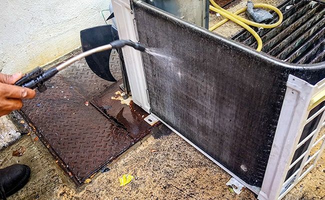 Person cleaning an outdoor air conditioner unit with a hose, removing dirt and debris.