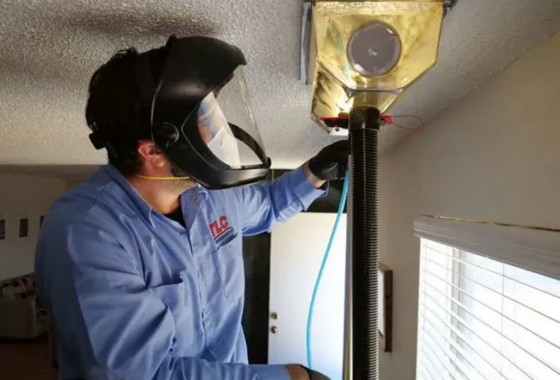 A person in protective gear inspects a duct from a ceiling. They wear a face shield and blue shirt, in a room with a window.