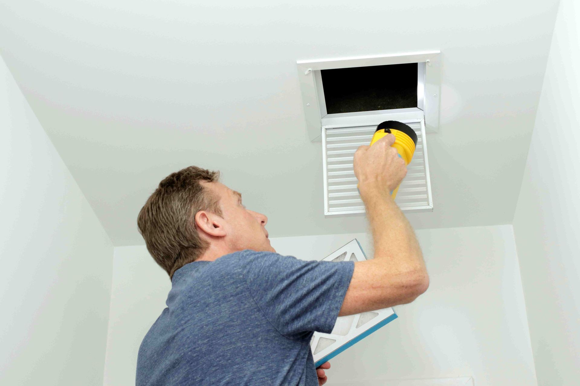 A man inspects an air vent filter with a flashlight in a white-walled room. He holds the filter in one hand.