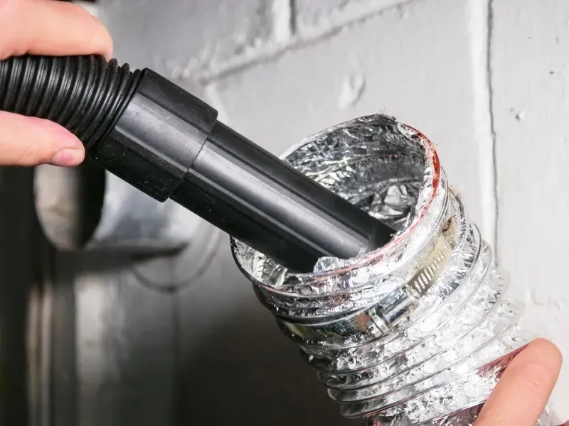 A person cleaning a dryer vent with a vacuum hose, removing lint from the metallic tubing attached to a white wall.