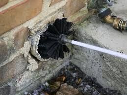 A person using a brush attached to a pole to clean out a dryer vent opening in brick.
