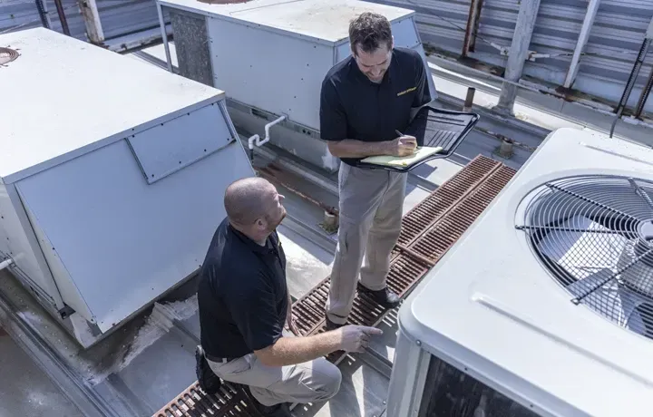 Two men inspecting HVAC units on a rooftop. One points while the other takes notes. Both wear black shirts and khakis.