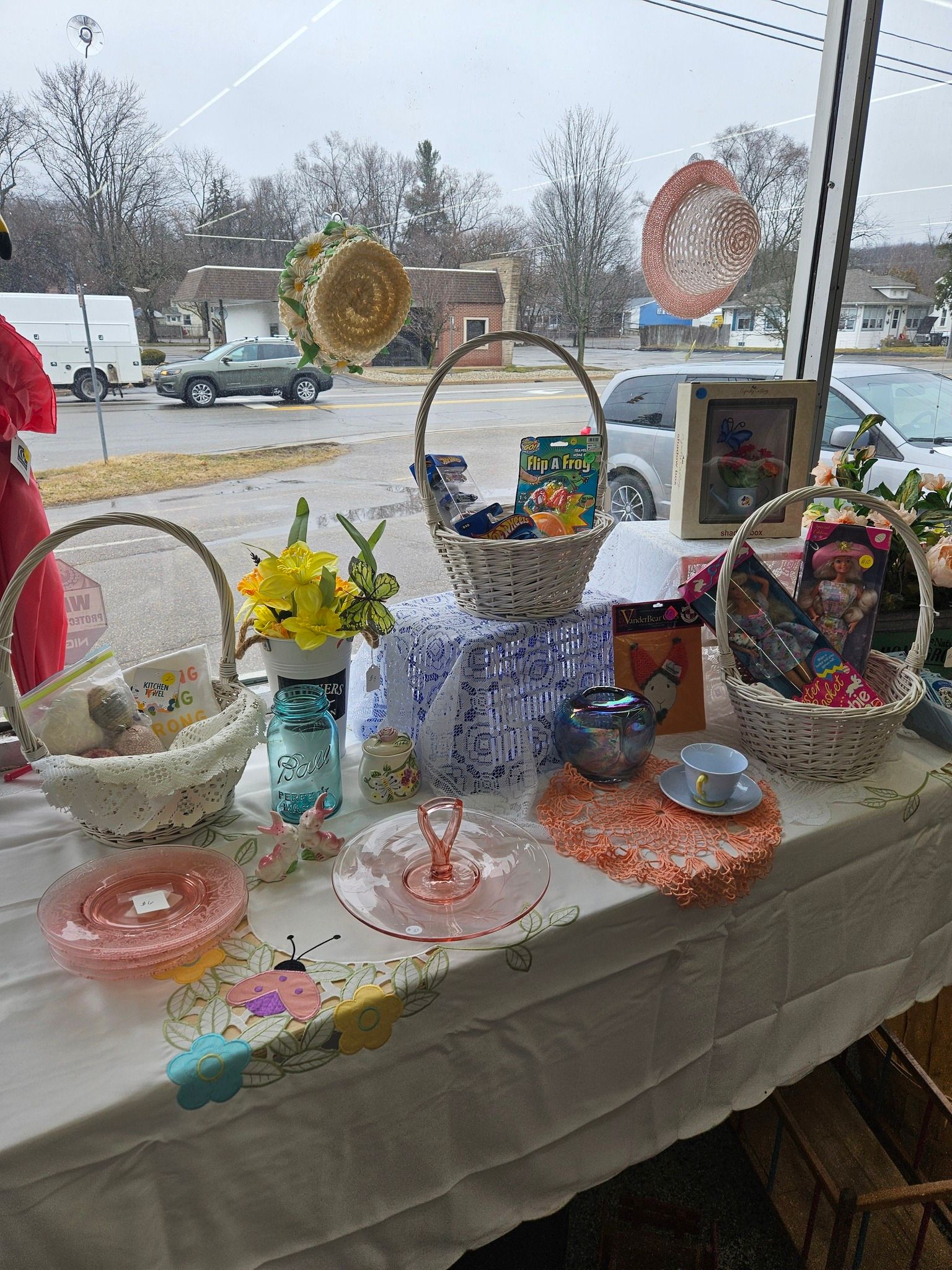 Table display of pastel handmade items and flowers in a shop window, with baskets, bowls, and gift boxes
