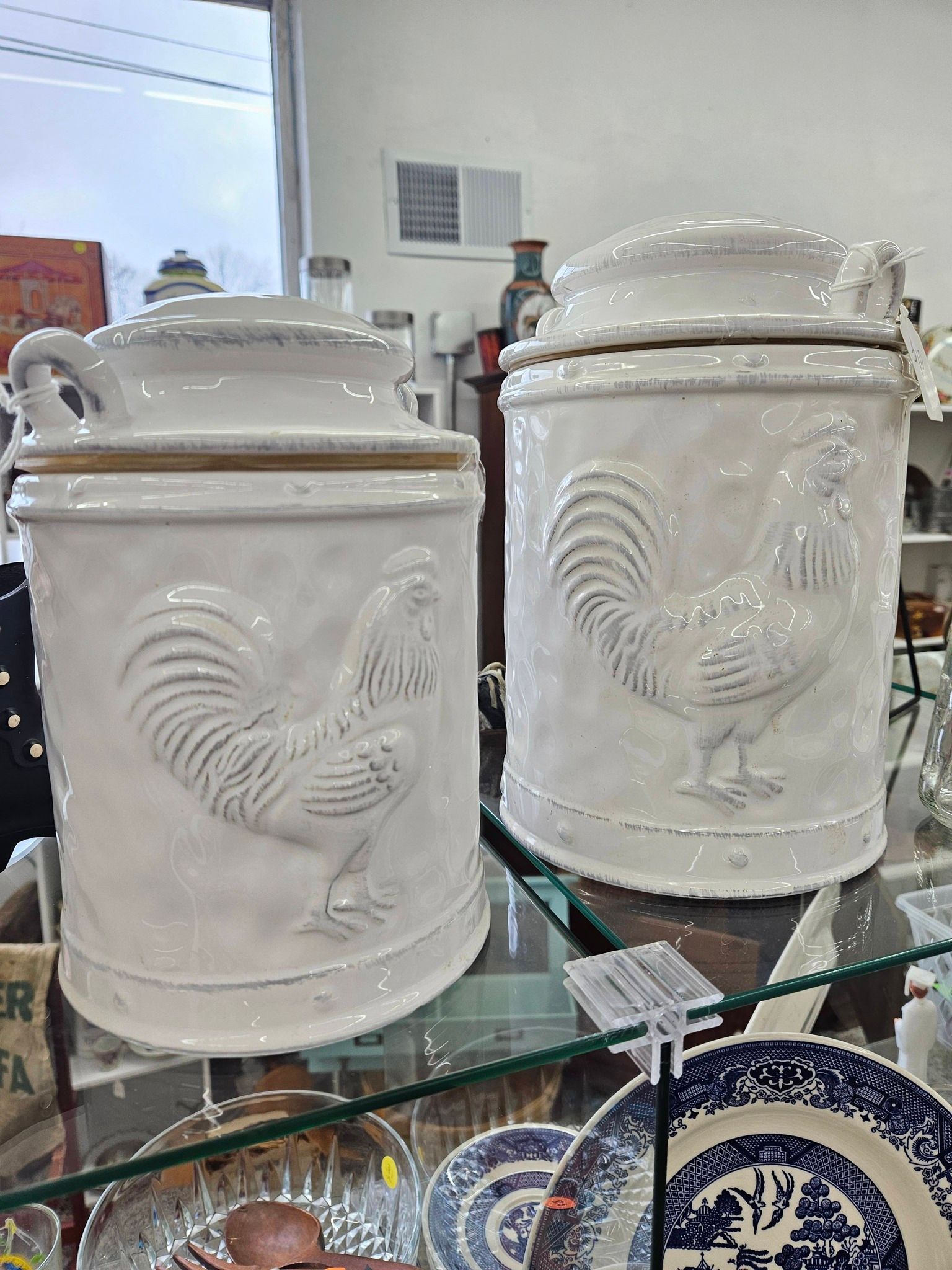 Two white ceramic rooster canisters with lids on a glass shelf in a store display