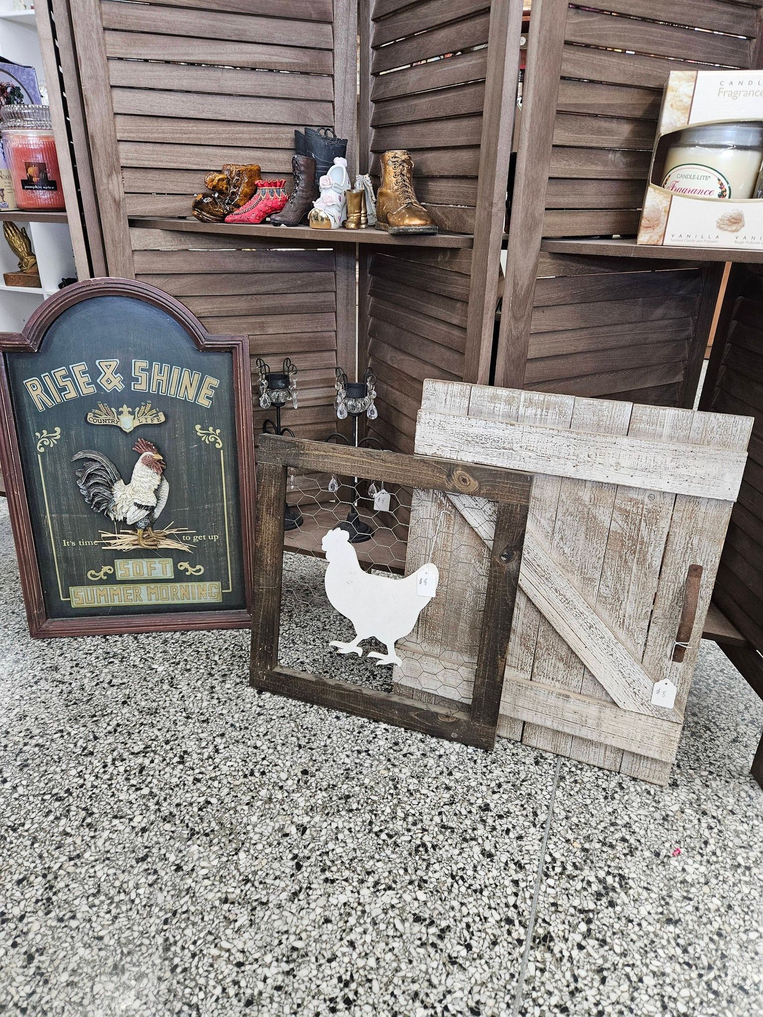 Rustic display with painted rooster sign, small figurines, and wooden crates on a gravel surface.