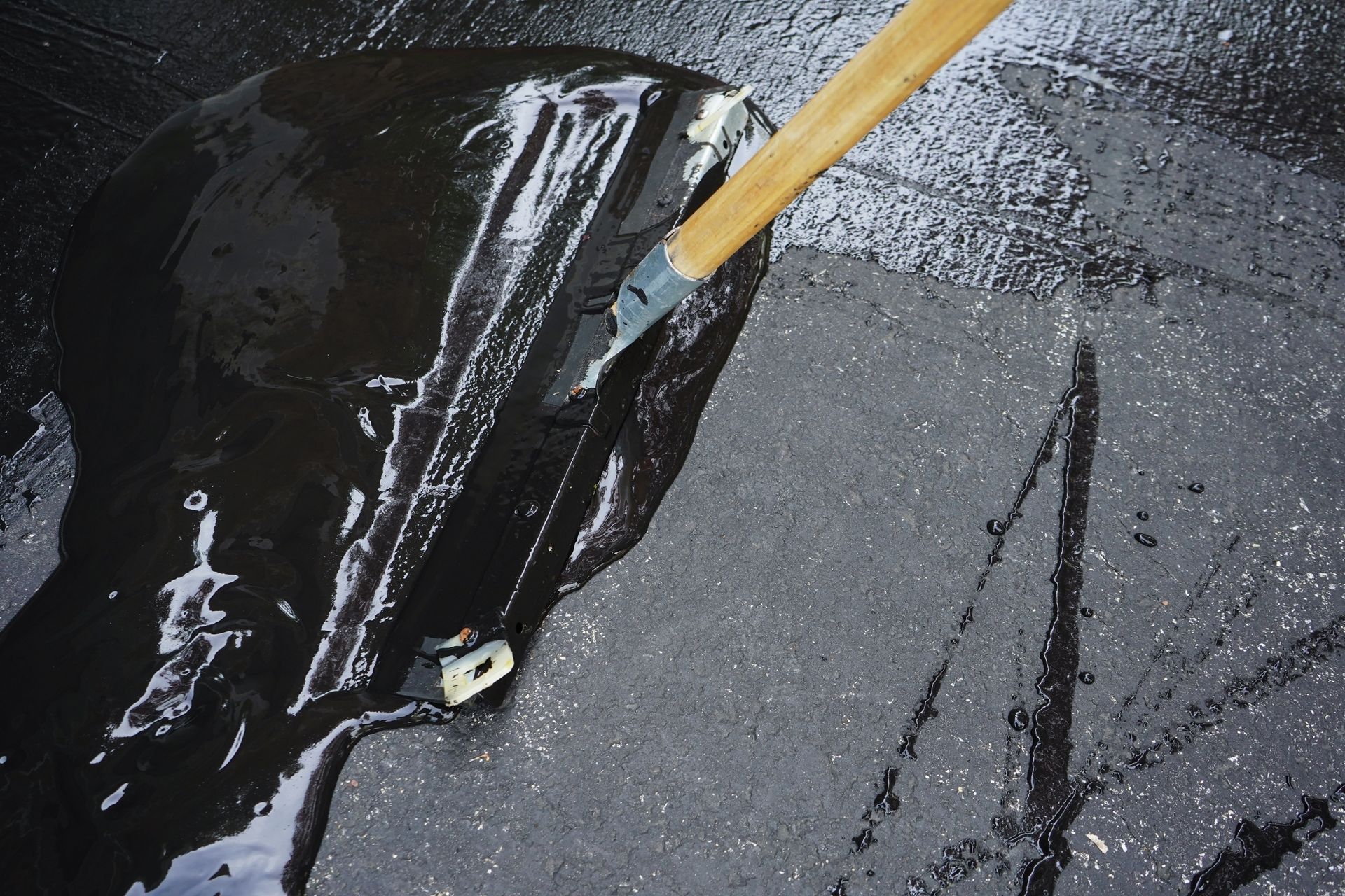 Road paving machine laying fresh black asphalt on a street beside a worker's boots Black liquid being poured onto a rough gray surface with a wooden stick stirring it