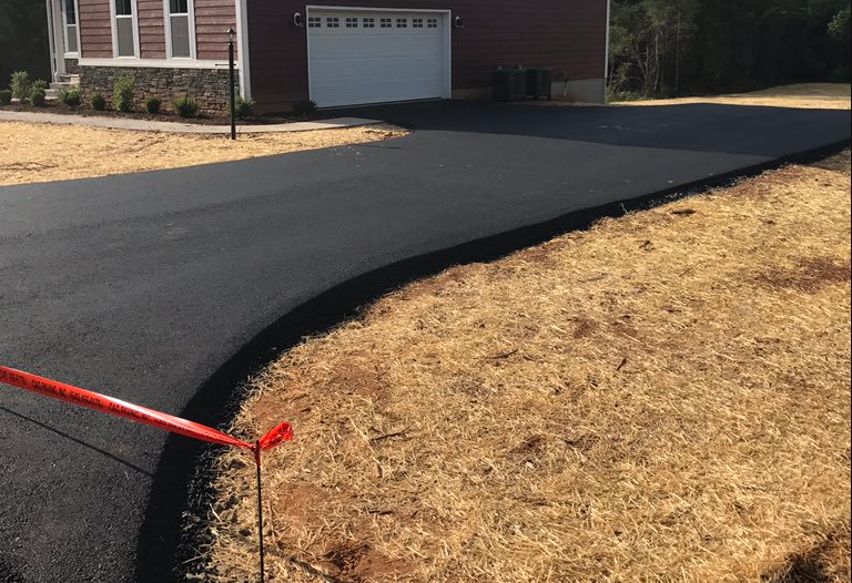 Fresh black asphalt driveway with a curved edge beside a brown house and dry grass, marked by a red stake