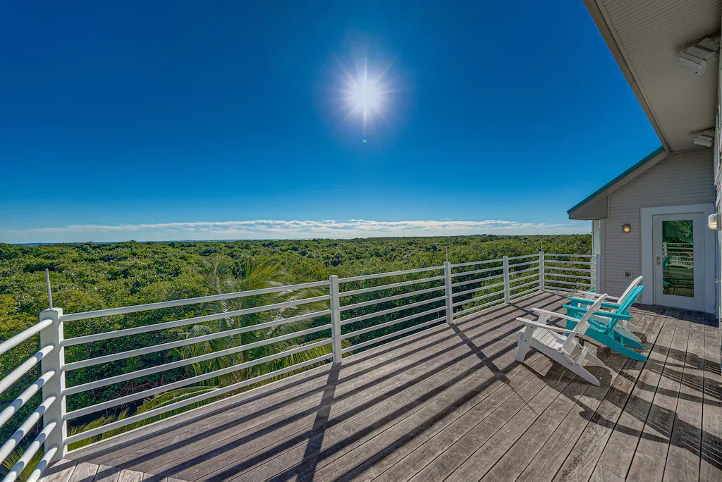 Deck with two chairs overlooking a dense green forest under a bright sun and blue sky.