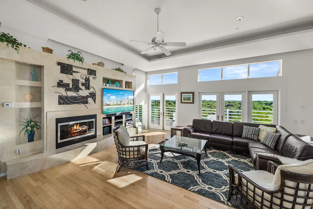Spacious living room with fireplace, TV, dark leather sectional, and large windows overlooking a lush green view.