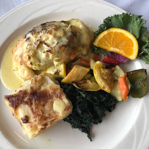 Plate with a baked dish in sauce, roasted vegetables, greens, and orange slice.
