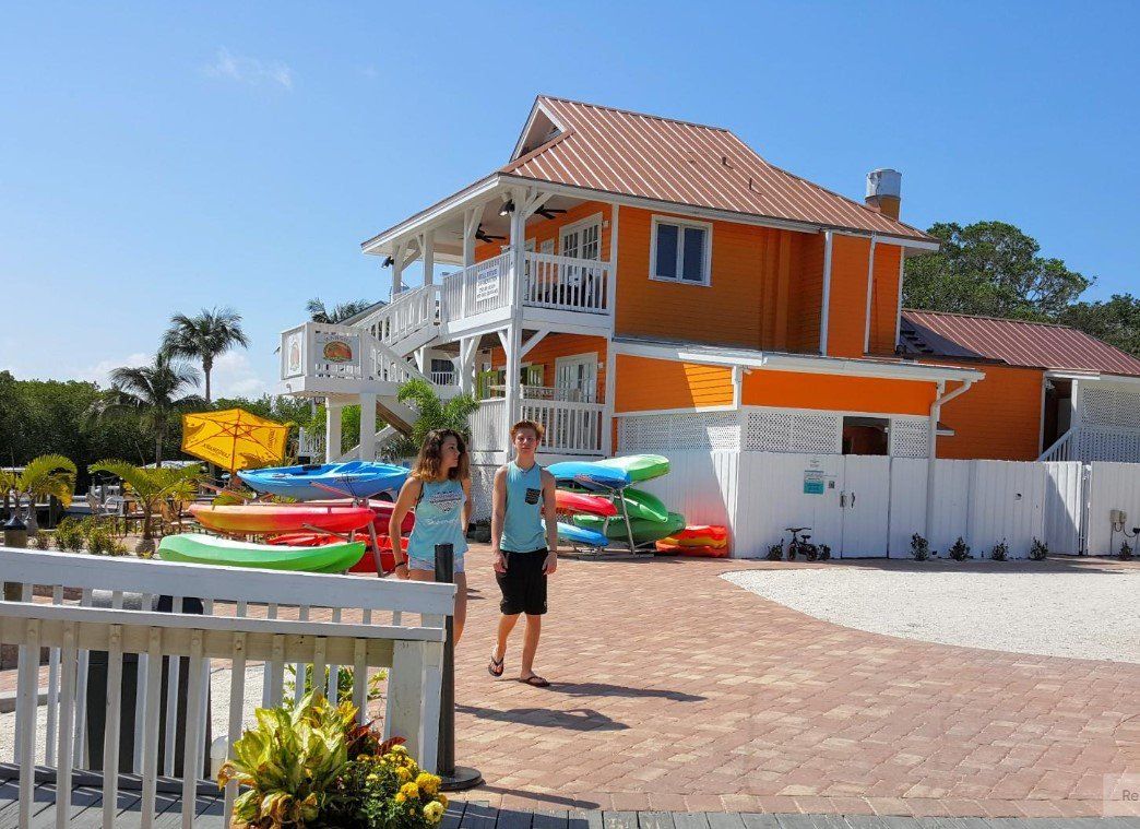 Two people walk toward an orange building. Kayaks are stacked nearby. Sunny day.