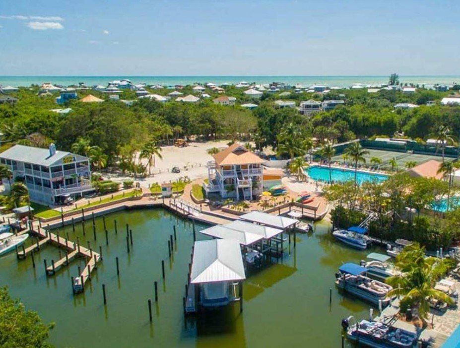 Aerial view of a waterfront property with a dock, boats, pool, and beach. Blue water and sky, green trees.