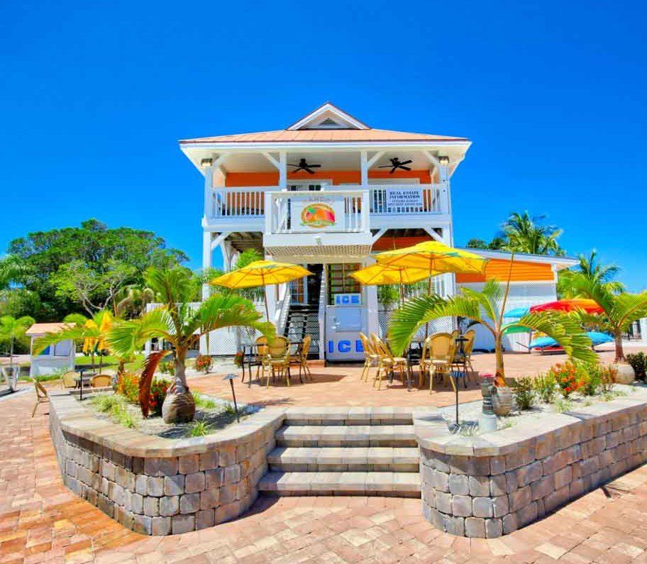 Two-story tropical restaurant with orange accents, yellow umbrellas, and palm trees; sunny day, beachfront setting.