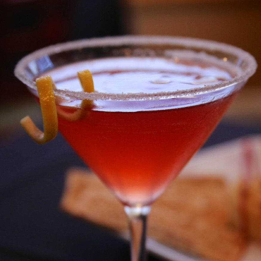 Cocktail in a martini glass with a sugar rim and lemon twist garnish. Red liquid.