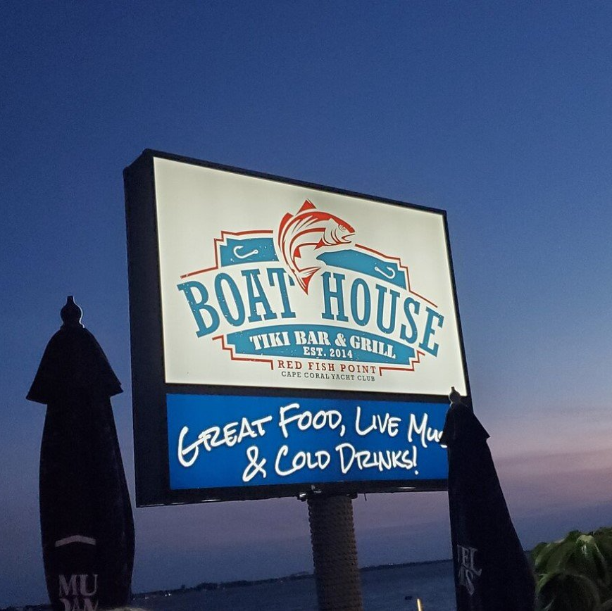 Sign for Boat House Tiki Bar & Grill with fish logo, blue and white text, night setting.
