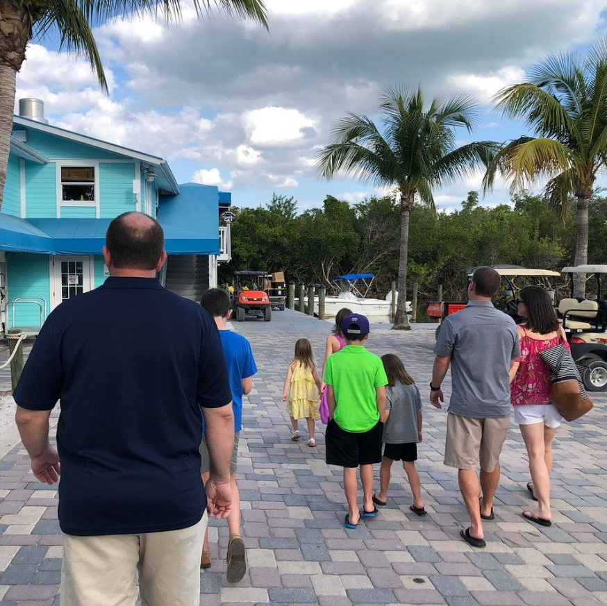 Families walking on a brick path towards a blue building with palm trees; golf carts are parked nearby.