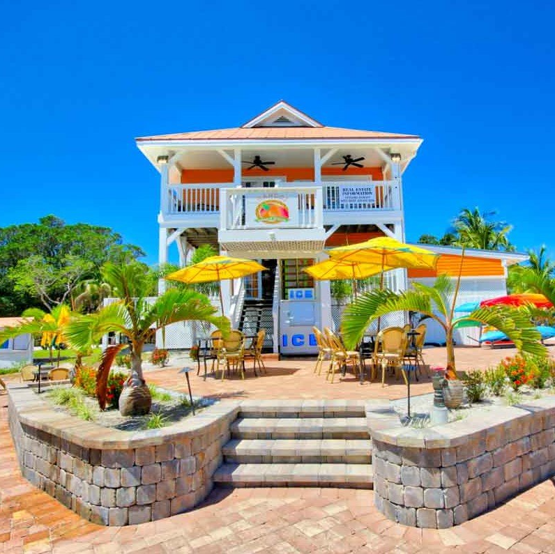 Tropical cafe with yellow umbrellas, an orange balcony, and palm trees against a blue sky.