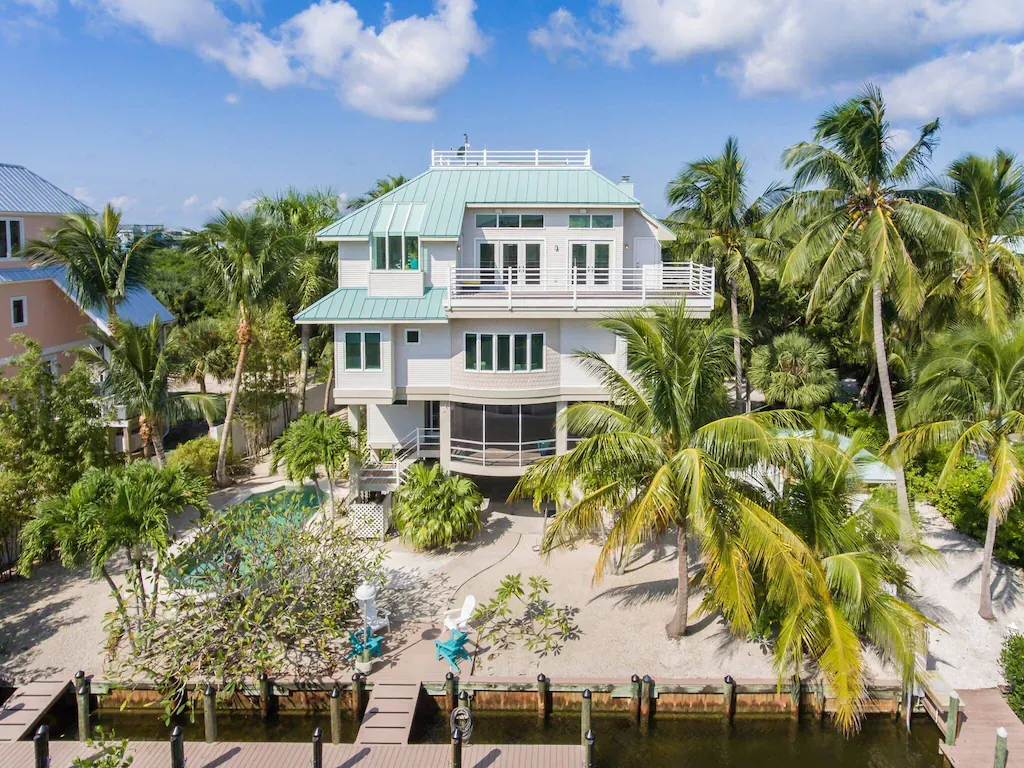 Tropical house with a turquoise roof and dock on a canal, surrounded by palm trees.