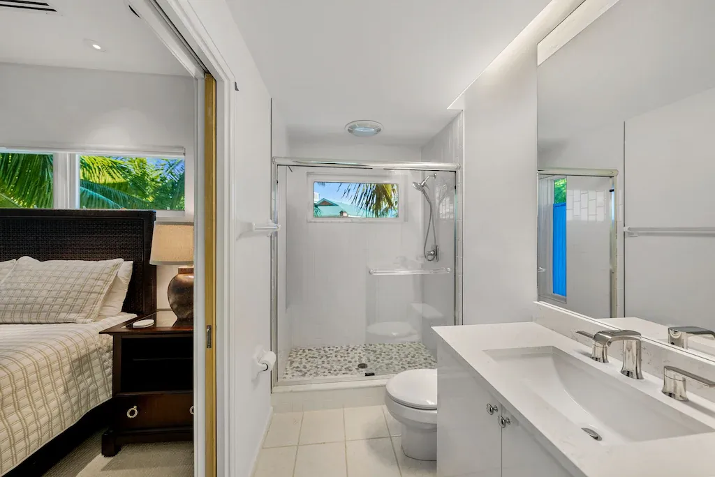 Bright, modern bathroom with a toilet, shower, and vanity, opening to a bedroom with a bed.