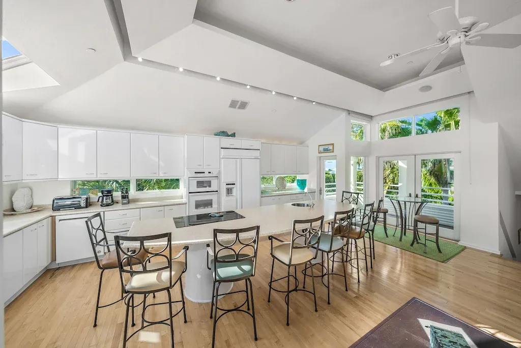 Bright kitchen with a large island, bar stools, and a dining area. Tall windows and skylights provide ample light.