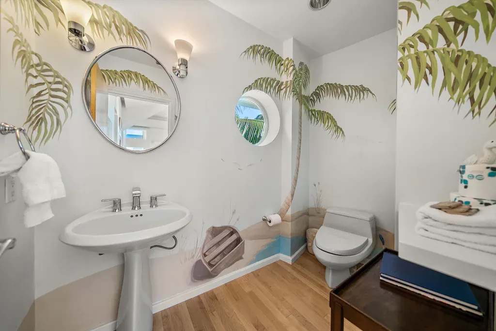 Tropical themed bathroom with palm tree mural, round mirror, pedestal sink, and porthole window.