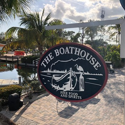 The Boathouse restaurant sign, circular, dark green with white lettering and boat illustration; palm trees and water in background.