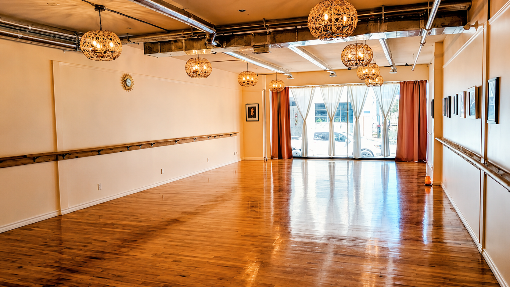 Empty wooden dance studio with polished floor, white walls, hanging lights, and large windows at the far end