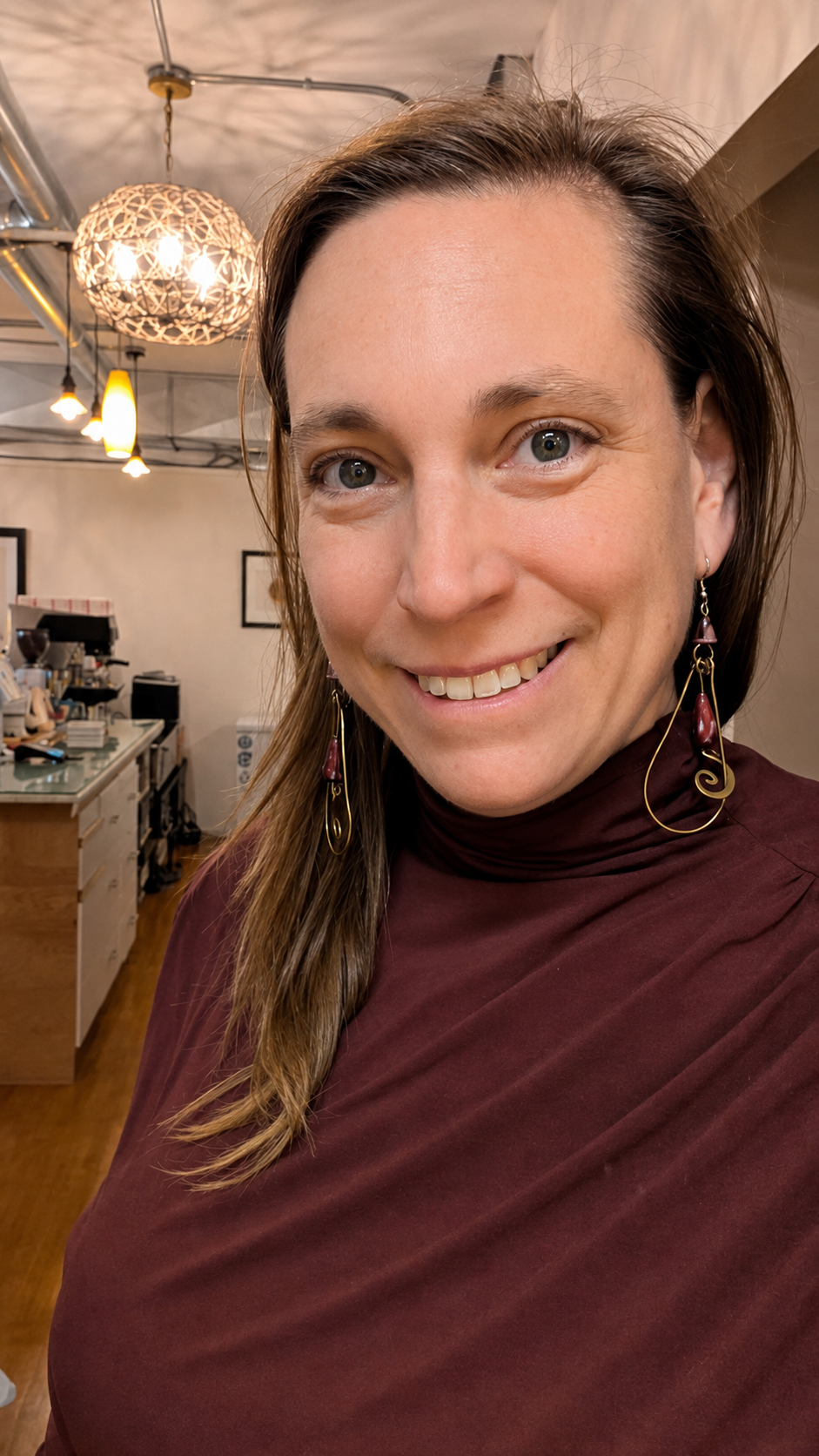 Smiling person in a maroon top indoors, with hanging lights and a warm café-like background