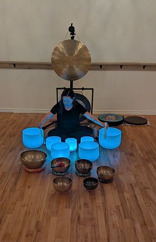 Person seated on floor amid glowing blue singing bowls with a large gong behind them