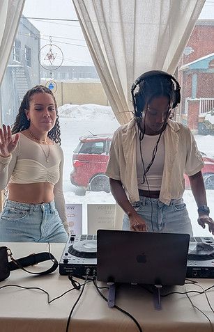 Two people standing at a laptop in front of a bright window with audio gear on the table
