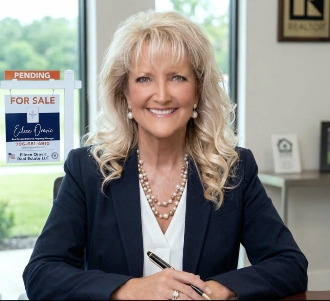 A professional realtor in a navy blazer sits at a desk with a pen, posing in front of a