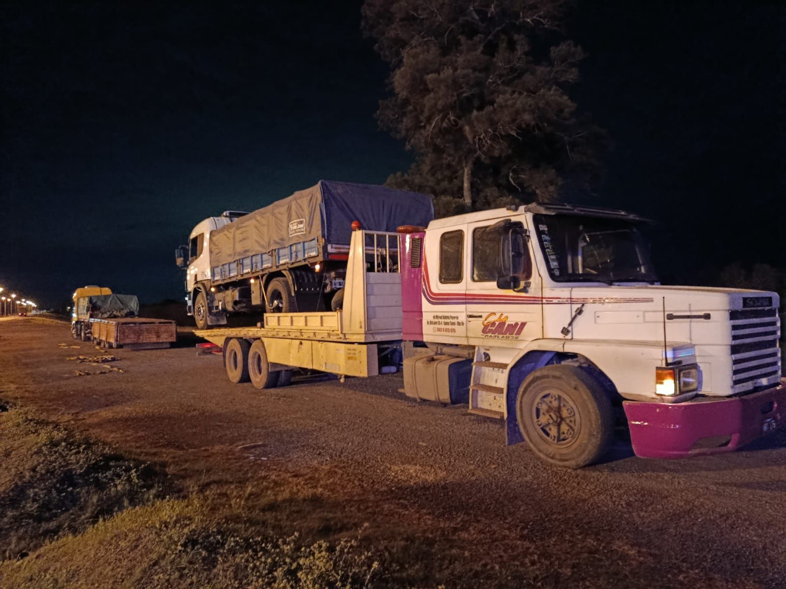 Un camión semirremolque blanco y magenta transporta carga al costado de la carretera por la noche.