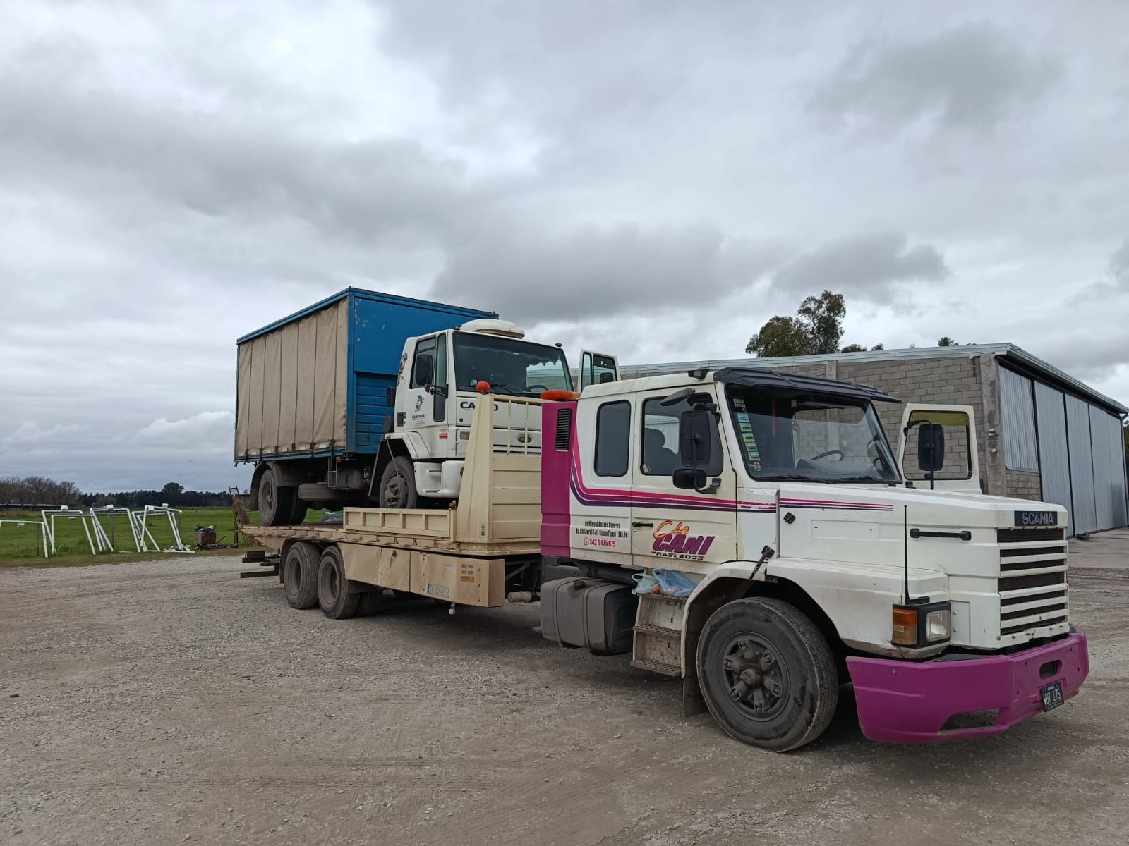 Camión de plataforma rosa y blanco transportando un contenedor azul en un terreno de grava bajo un cielo nublado.