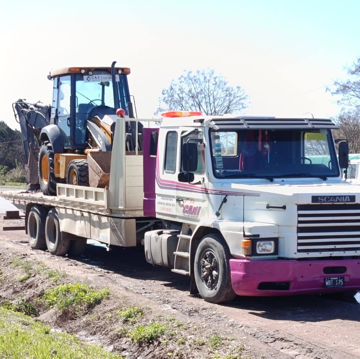Una grúa transporta un tractor azul en una plataforma junto a un camino de tierra bajo la luz del sol.