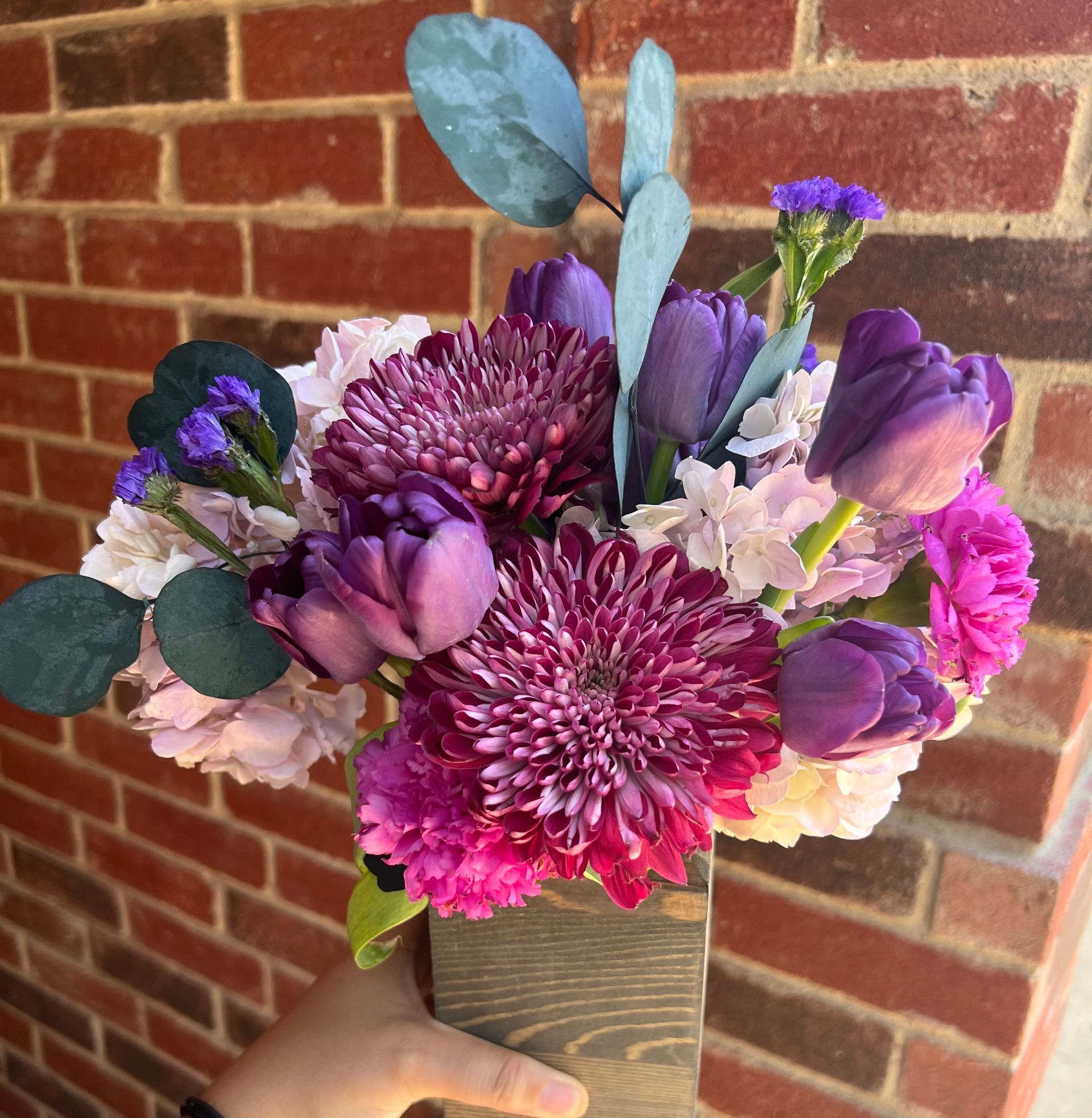 A person is holding a vase of purple flowers in front of a brick wall