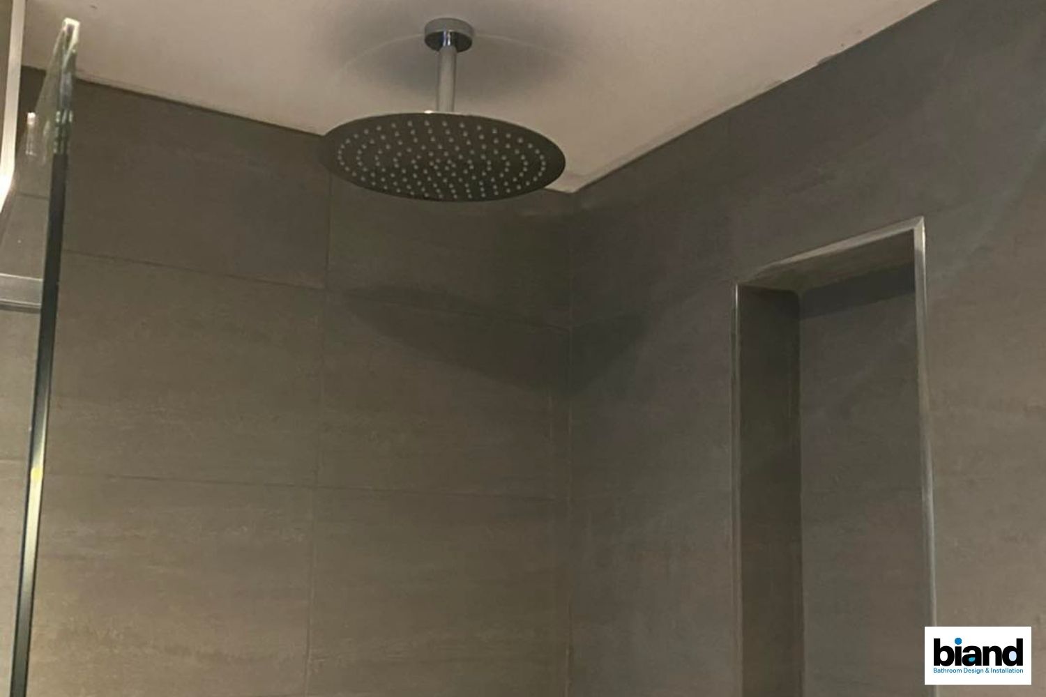 Shower with a large, round rain showerhead mounted on the ceiling. Gray tiled walls and a recessed niche.