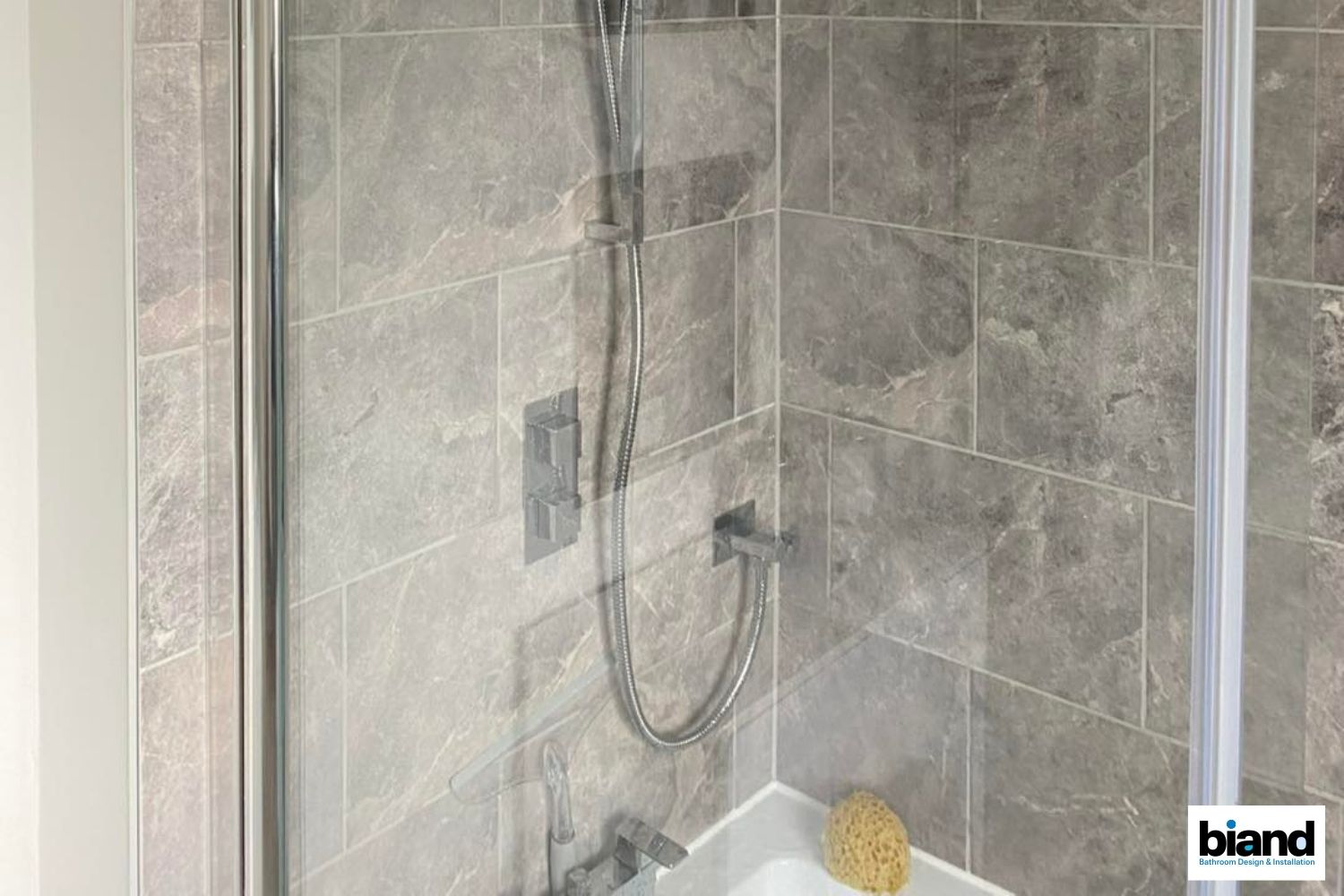 Shower enclosure with gray tiled walls, glass door, showerhead, and soap sponge.