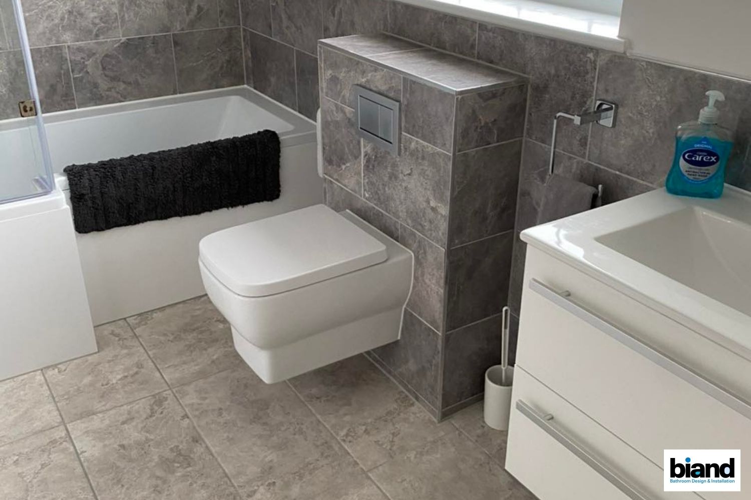 Modern gray tiled bathroom with floating toilet, vanity, and bathtub.